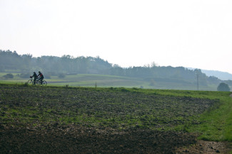 Zwei Radfahrende hinter einem frisch geflügtem Acker vor einem Hügel enlangradelnd