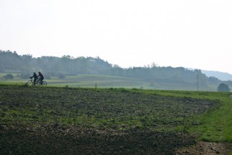 Zwei Radfahrende hinter einem frisch geflügtem Acker vor einem Hügel enlangradelnd