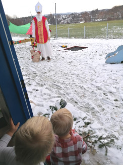 Foto vom Hl. Nikolaus im Garten mitten im Schnee mit 2 Kindern, die aus der Tür der KiTa EschenauNeu spitzen